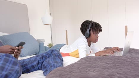Afroamerikanischer-Vater-Und-Sohn-Liegen-Zu-Hause-Mit-Laptop-Und-Smartphone-Im-Bett,-Zeitlupe