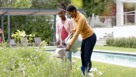 Happy-african-american-daughter-and-mother-watering-plants-in-sunny-garden,-slow-motion