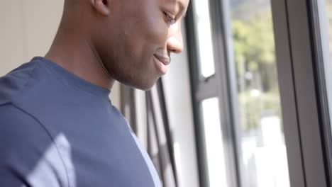 Hombre-Afroamericano-Usando-Un-Teléfono-Inteligente-Y-Mirando-Por-La-Ventana-En-Casa,-Cámara-Lenta