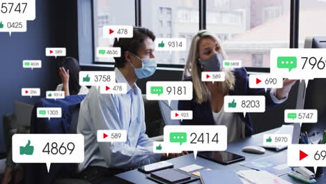 Animation-of-social-media-icons-over-diverse-man-and-woman-wearing-face-masks-discussing-at-office
