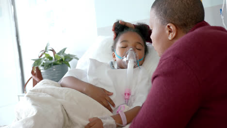 African-american-woman-with-face-mask-stroking-daughter-with-oxygen-mask-in-hospital,-slow-motion