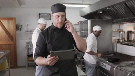 Chef-Masculino-Caucásico-Usando-Tableta-Y-Hablando-Por-Teléfono-Inteligente-En-La-Cocina,-Cámara-Lenta