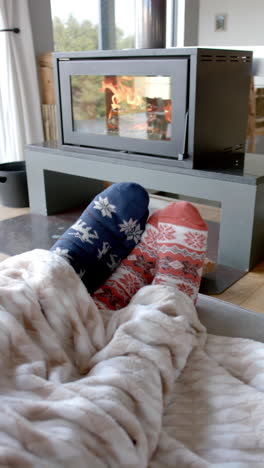 Vertical-video-of-feet-of-biracial-couple-sitting-on-sofa-in-front-of-fireplace-at-home,-slow-motion