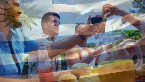 Composite-Video-Der-Uruguayischen-Flagge-über-Verschiedenen-Freunden,-Die-Beim-Mittagessen-Im-Park-Mit-Getränken-Anstoßen