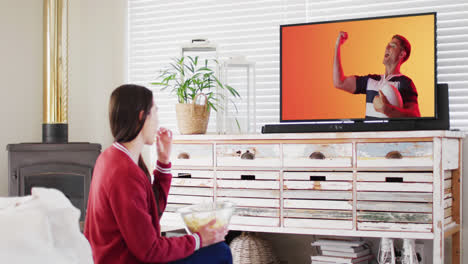Caucasian-woman-watching-tv-with-caucasian-male-rugby-player-with-ball-on-screen