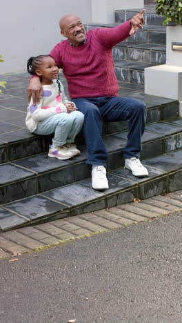 Happy-african-american-grandfather-sitting-on-steps-and-embracing-granddaughter,-slow-motion