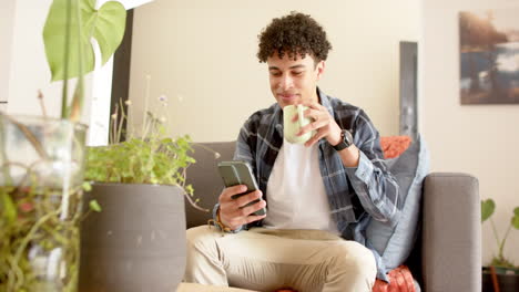 Biracial-man-sitting-on-couch,-using-smartphone-and-drinking-coffee-at-home,-slow-motion
