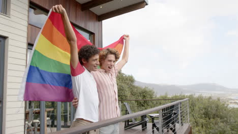 Feliz-Pareja-Gay-Diversa-Sosteniendo-La-Bandera-Lgbt-Del-Arco-Iris-En-El-Balcón-De-Casa,-Cámara-Lenta