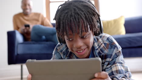 Glücklicher-Afroamerikanischer-Junge-Mit-Kopfhörern-Und-Tablet,-Das-Zu-Hause-Auf-Dem-Boden-Liegt,-Zeitlupe