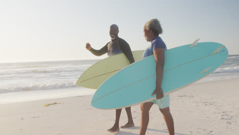 Glückliches-älteres-Afroamerikanisches-Paar,-Das-In-Zeitlupe-Am-Strand-Spazieren-Geht-Und-Surfbretter-Hält