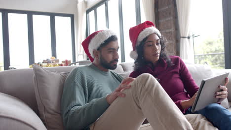 Biracial-couple-wearing-santa-claus-hats-sitting-on-sofa-using-tablet-at-home,-slow-motion