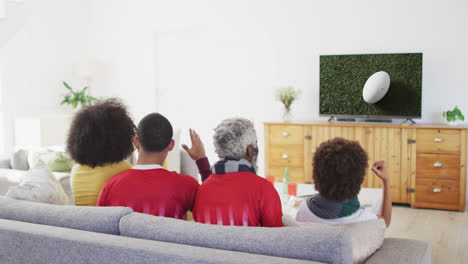 Biracial-family-watching-tv-with-rugby-ball-at-stadium-on-screen