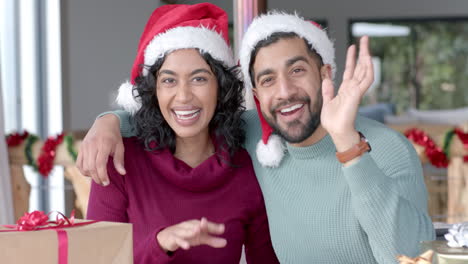 Happy-biracial-couple-wearing-santa-claus-hats-having-christmas-video-call-at-home,-in-slow-motion