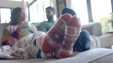 Happy-biracial-couple-on-sofa-in-warm-socks-under-blanket-drinking-coffee-at-home,-slow-motion