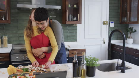 Happy-caucasian-lesbian-couple-preparing-food,-embracing-and-using-tablet-in-kitchen,-copy-space