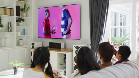 Asian-family-watching-tv-with-diverse-male-rugby-players-with-ball-on-screen