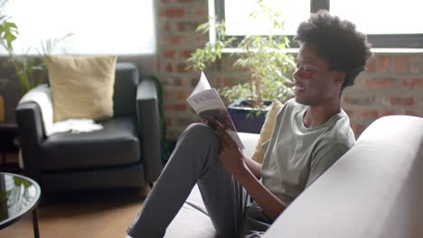 African-american-man-with-under-eye-patches-reading-book-on-sofa-at-home,-slow-motion