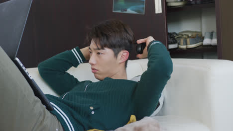 Asiatischer-Junge-Mit-Kopfhörern-Und-Laptop,-Der-Zu-Hause-Auf-Der-Couch-Liegt
