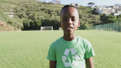 Un-Niño-Afroamericano-Con-Una-Camiseta-Verde-De-Reciclaje-Se-Encuentra-En-Un-Soleado-Campo-De-Fútbol,-Con-Copia-