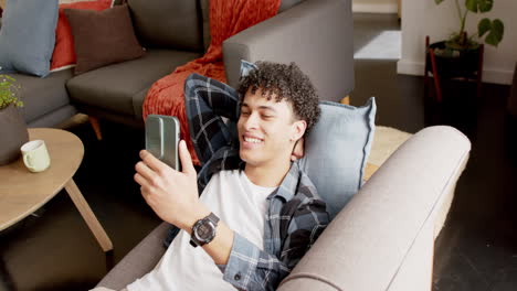Biracial-man-lying-on-couch-and-holding-smartphone-at-home,-slow-motion