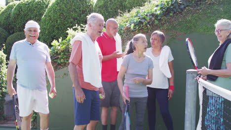 Diverse-group-of-happy-male-and-female-seniors-talking-at-sunny-outdoor-tennis-court,-slow-motion
