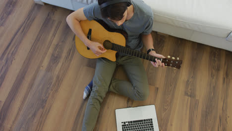 Overhead-view-of-asian-boy-wearing-headphones-playing-guitar-looking-at-the-laptop-at-home