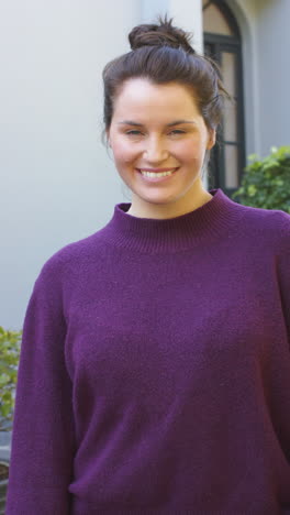 Vertical-video-of-happy-caucasian-woman-in-purple-sweater-smiling-in-front-of-house-in-garden