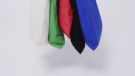 A-collection-of-colorful-tote-bags-in-white,-green,-red,-black,-and-blue-hangs-against-a-plain-backg