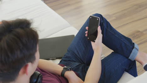 Glücklicher-Asiatischer-Männlicher-Teenager,-Der-Kopfhörer-Trägt-Und-Im-Wohnzimmer-Sein-Smartphone-Benutzt