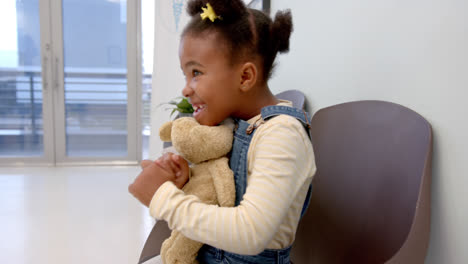 Happy-african-american-girl-sitting-in-hospital-waiting-room-and-holding-mascot,-slow-motion
