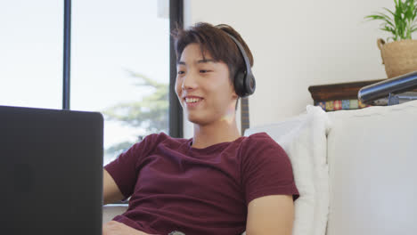 Glücklicher-Asiatischer-Männlicher-Teenager,-Der-Kopfhörer-Trägt-Und-Im-Wohnzimmer-Einen-Laptop-Benutzt