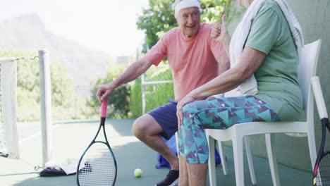 Happy-senior-caucasian-couple-taking-a-break,-talking-at-sunny-outdoor-tennis-court,-slow-motion