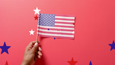 Animation-of-stars-falling-over-hand-holding-flag-of-united-states-of-america-on-red-background