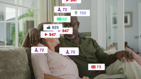 Animation-of-social-media-icons-and-numbers-over-senior-biracial-couple-at-home