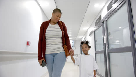 Afroamerikanische-Mutter-Und-Tochter-Im-Krankenhauskleid-Halten-Händchen-Beim-Gehen-Im-Flur,-Zeitlupe