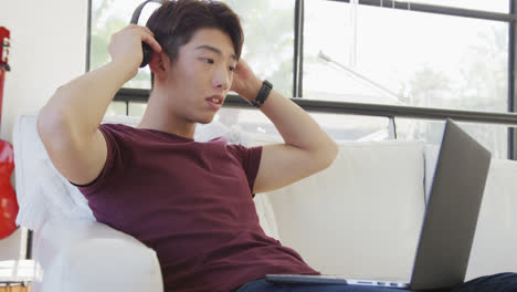 Asian-male-teenager-wearing-headphones-and-using-laptop-in-living-room