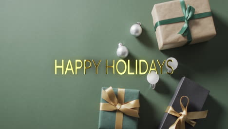 Happy-holidays-text-in-gold-over-christmas-baubles-and-gifts-on-green-background