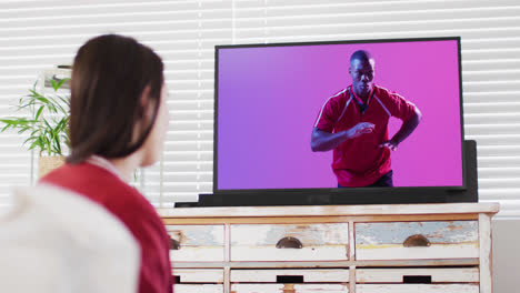 Caucasian-woman-watching-tv-with-african-american-male-rugby-player-catching-ball-on-screen