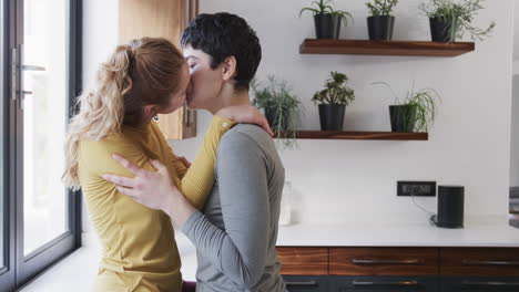 Happy-caucasian-lesbian-couple-embracing-and-kissing-in-sunny-kitchen,-copy-space