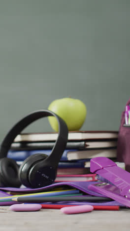 Vertical-video-of-headphones,-schoolbooks-and-apple-on-desk-with-chalk-board-in-background