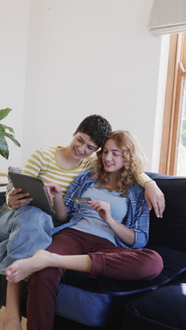 Glückliches-Kaukasisches-Lesbisches-Paar,-Das-Auf-Dem-Sofa-Sitzt,-Sich-Umarmt-Und-Ein-Tablet-Im-Sonnigen-Wohnzimmer-Benutzt