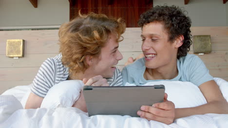 Happy-diverse-gay-male-couple-lying-on-bed-and-using-tablet-at-home,-slow-motion