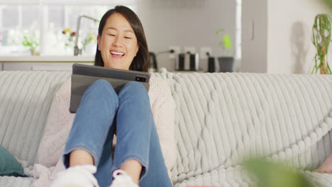 Video-Einer-Glücklichen-Asiatischen-Frau,-Die-Mit-Einem-Tablet-Auf-Dem-Sofa-Ruht