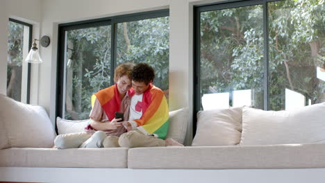 Diverse-gay-male-couple-with-rainbow-lgbt-flag,-using-smartphone-at-home,-slow-motion,-copy-space
