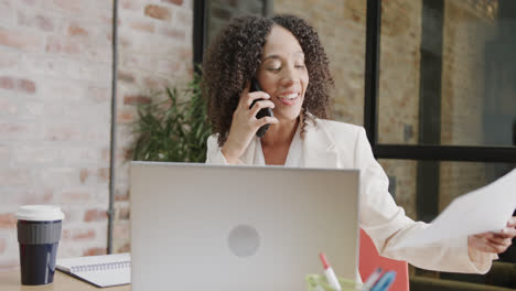 Happy-biracial-businesswoman-at-desk-with-laptop-holding-document-and-talking-on-phone,-slow-motion