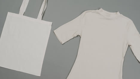 Close-up-of-white-bag-and-t-shirt-on-grey-background,-with-copy-space,-slow-motion