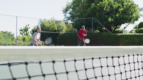 Diverse-senior-man-and-woman-playing-tennis-doubles-on-sunny-court,-slow-motion