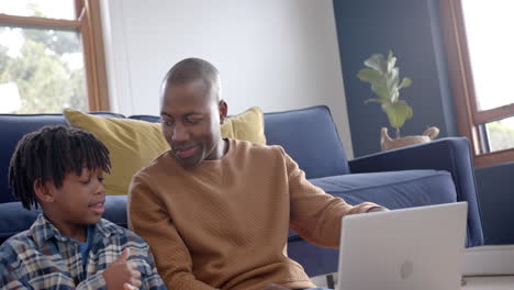 Happy-african-american-father-and-son-sitting-on-floor-and-using-laptop-at-home,-slow-motion