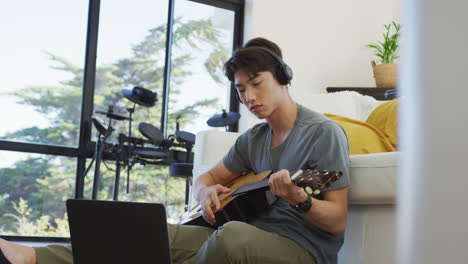 Asiatischer-Junge-Mit-Kopfhörern,-Der-Gitarre-Spielt-Und-Zu-Hause-Auf-Den-Laptop-Schaut
