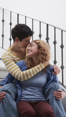 Happy-caucasian-lesbian-couple-sitting-on-stairs,-embracing-and-smiling-in-sunny-house